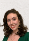 Portrait of Niamh Nolan smiling in front of a white textured wall, wearing a green top with gold earrings and layered necklaces.